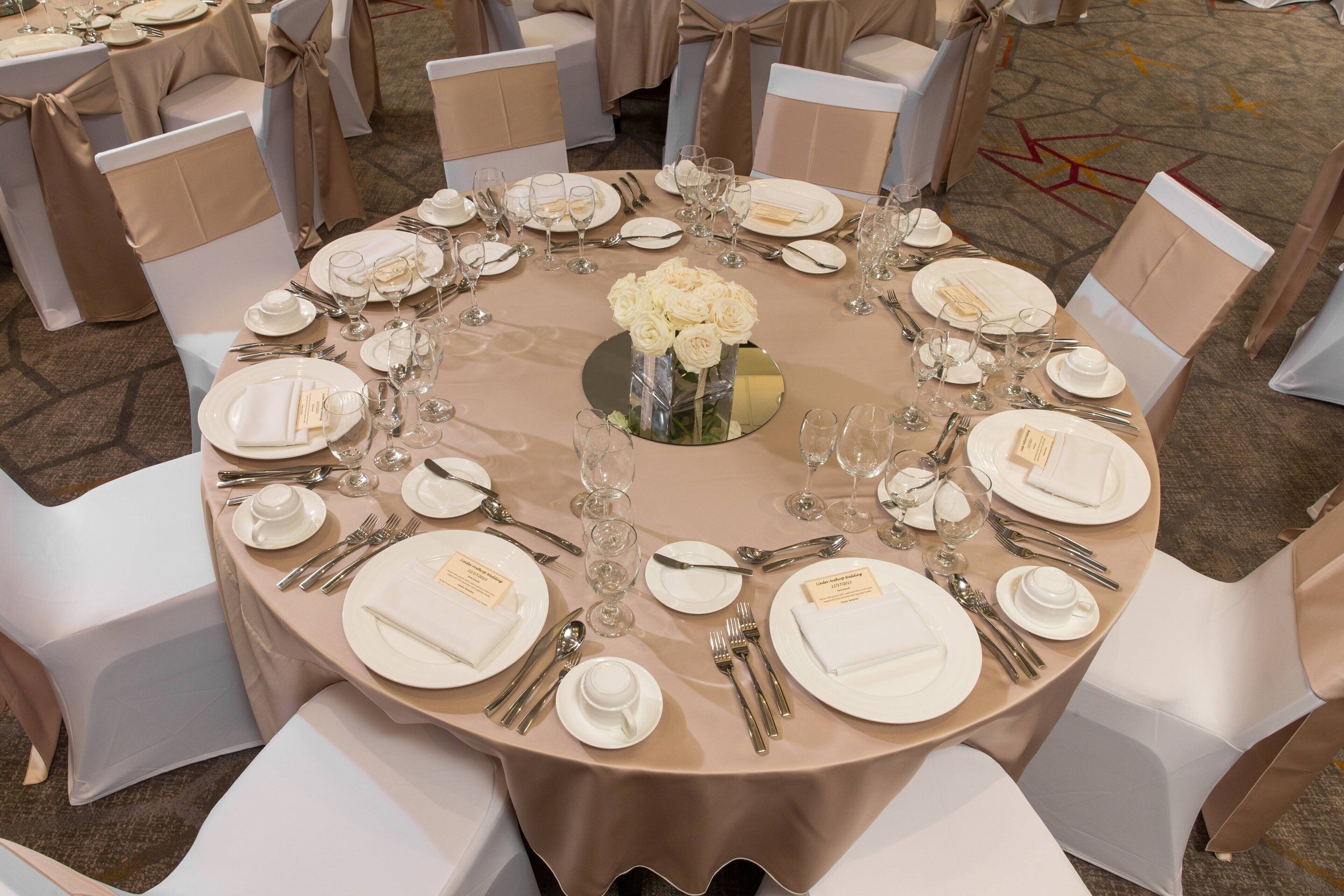 A round table set for a formal event with white chairs, a beige tablecloth, and a central floral arrangement.