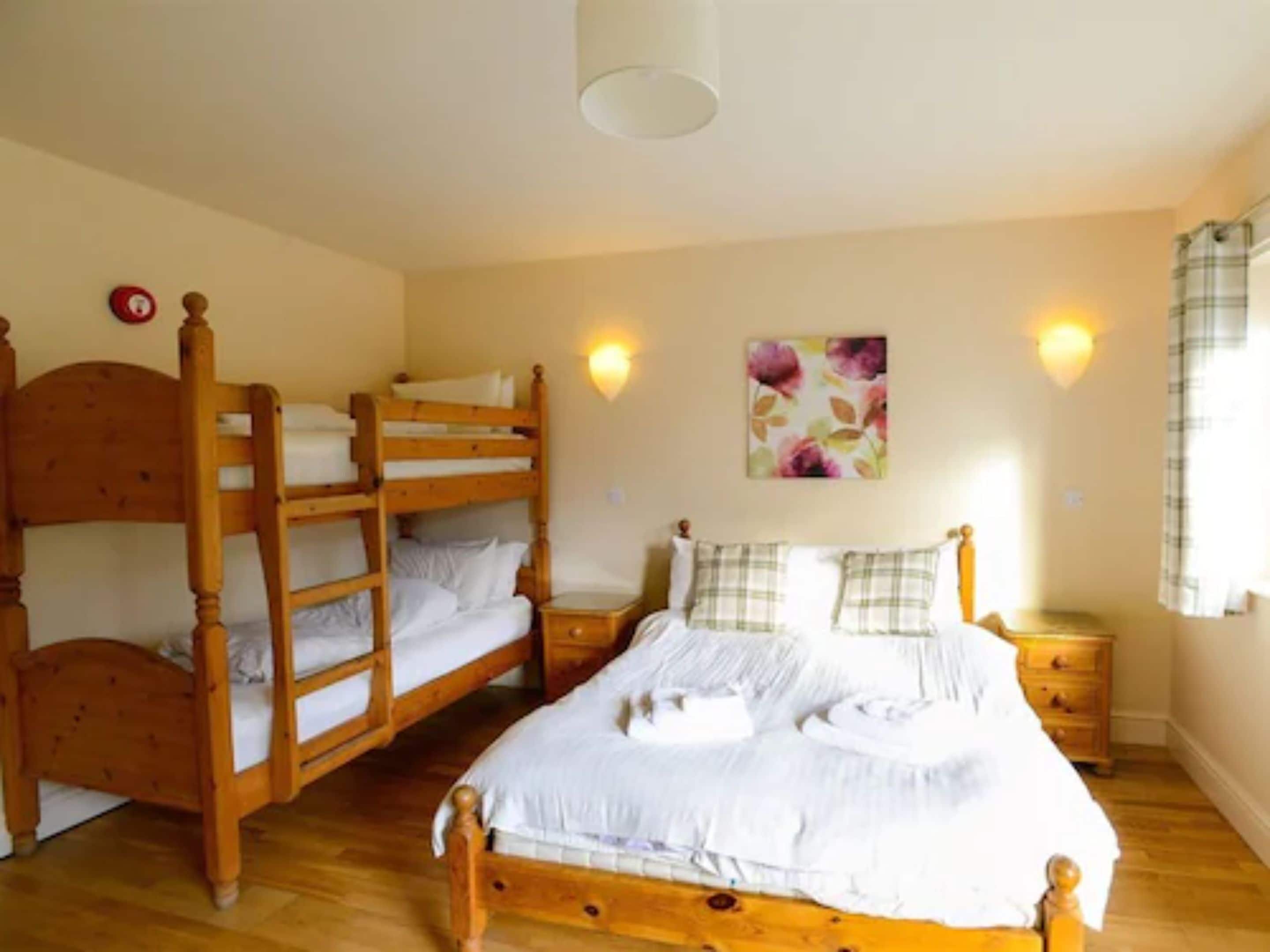 A bedroom with a bunk bed, a single bed, wooden headboards, a nightstand, a wall-mounted light, and a floral painting.