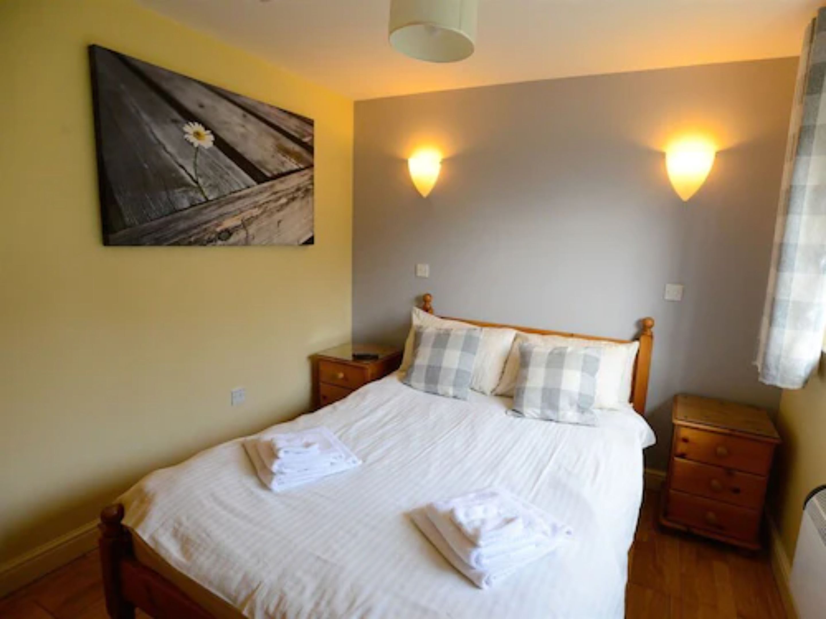 A neatly made bed with white linens and folded towels, two bedside tables, a wall-mounted artwork, and two wall sconces.