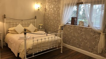 Un dormitorio ordenado con cama, mesitas de noche, un ventanal con cortinas y un papel tapiz floral en la pared.