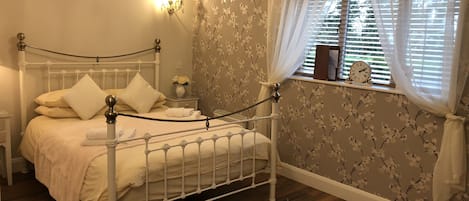 A neatly arranged bedroom with a bed, bedside tables, a nightstand, a window with curtains, and a wall with floral wallpaper.