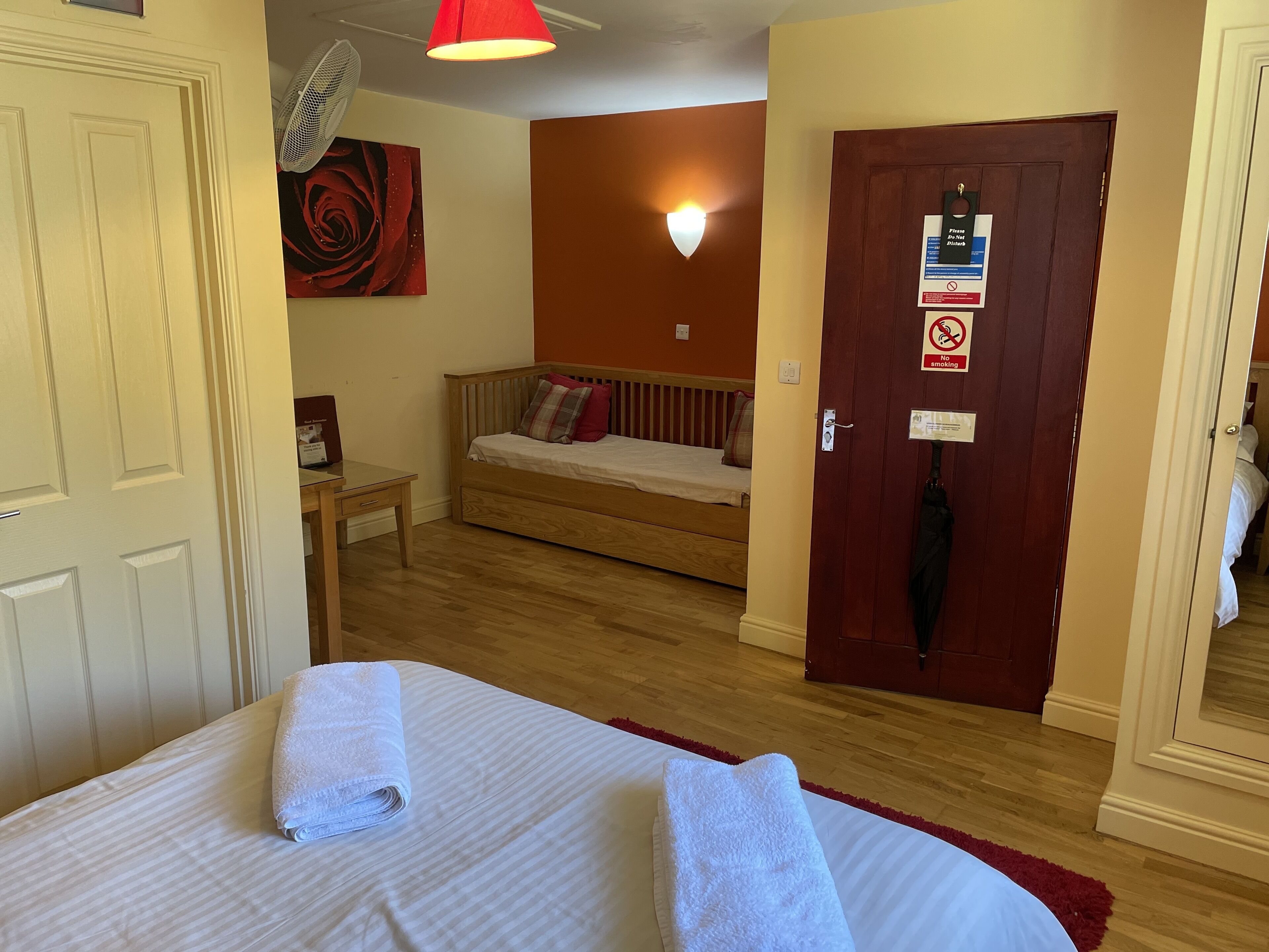 Habitación de hotel con cama, una mesita, un ventilador de pared, una puerta roja con un letrero y una pared roja con una pintura de rosa.