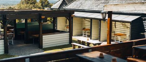 A covered outdoor seating area with wooden tables and benches, a pergola structure, and a view of the surrounding landscape.