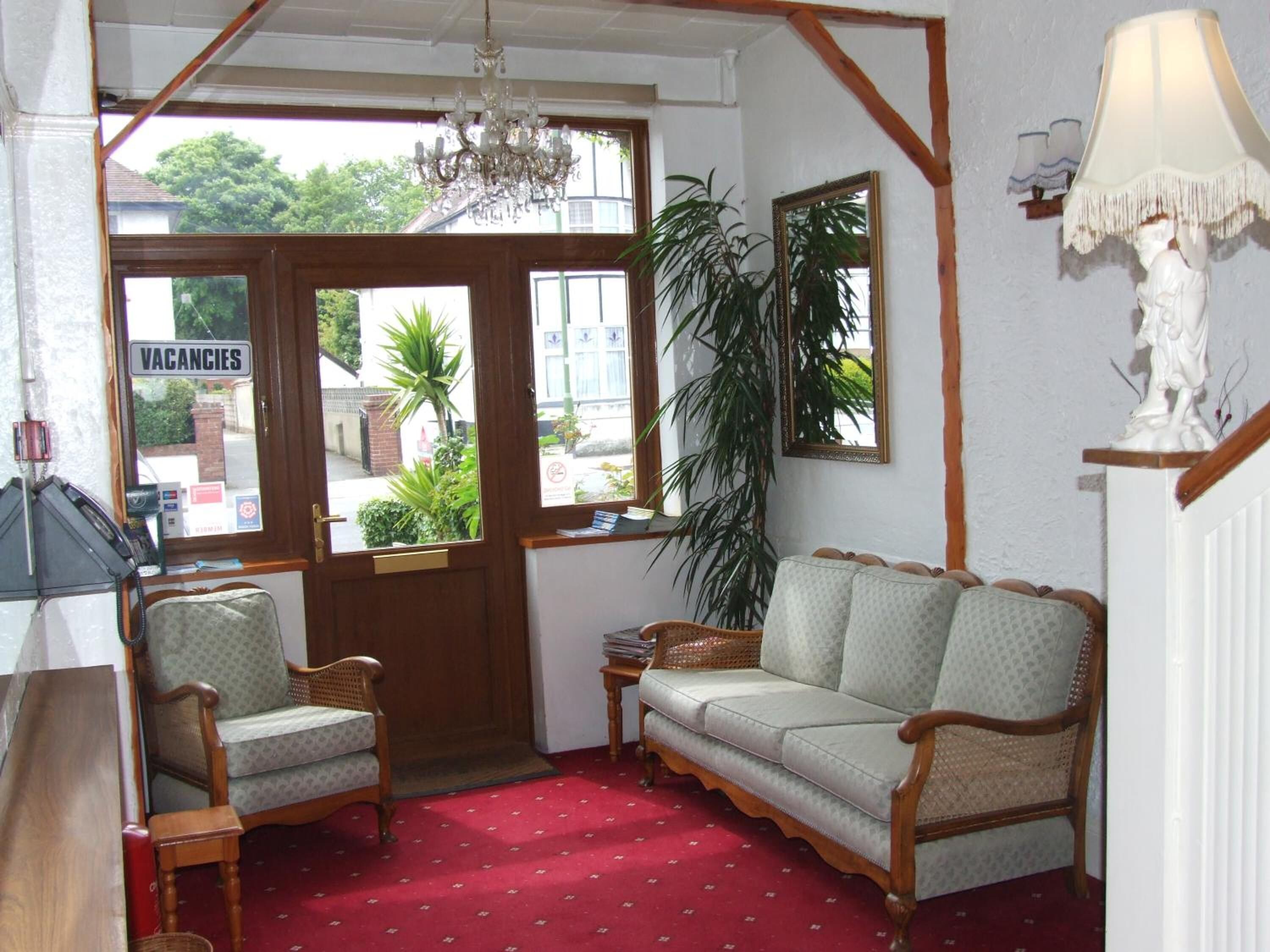 A room with a red carpet, wooden furniture, a chandelier, a large window with a 'VACANCIES' sign, and a plant. - The Braedene Lodge B&B (Paignton)
