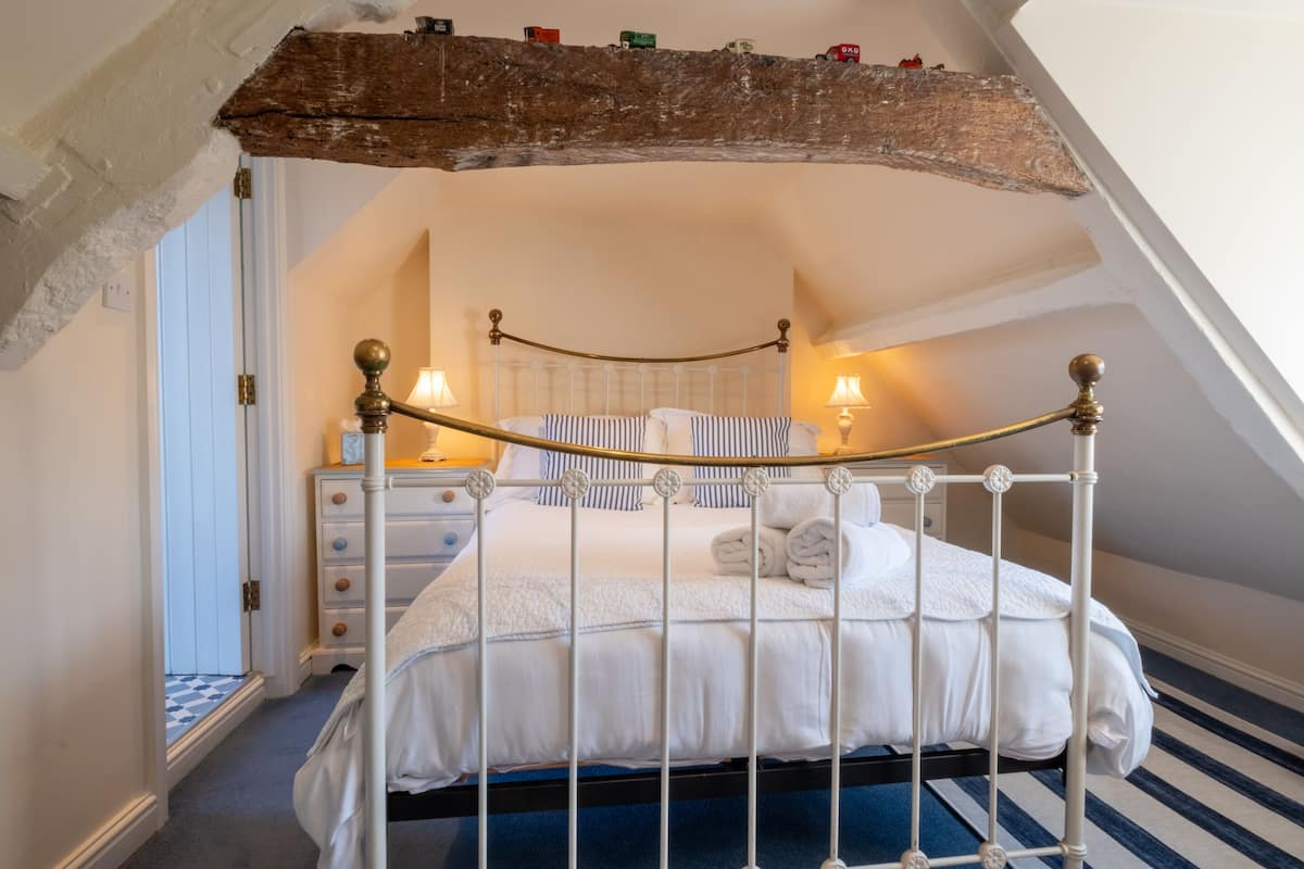 A four-poster bed with white bedding and a blue and white striped pillow. There is a wooden beam above the bed and a white door to the left.