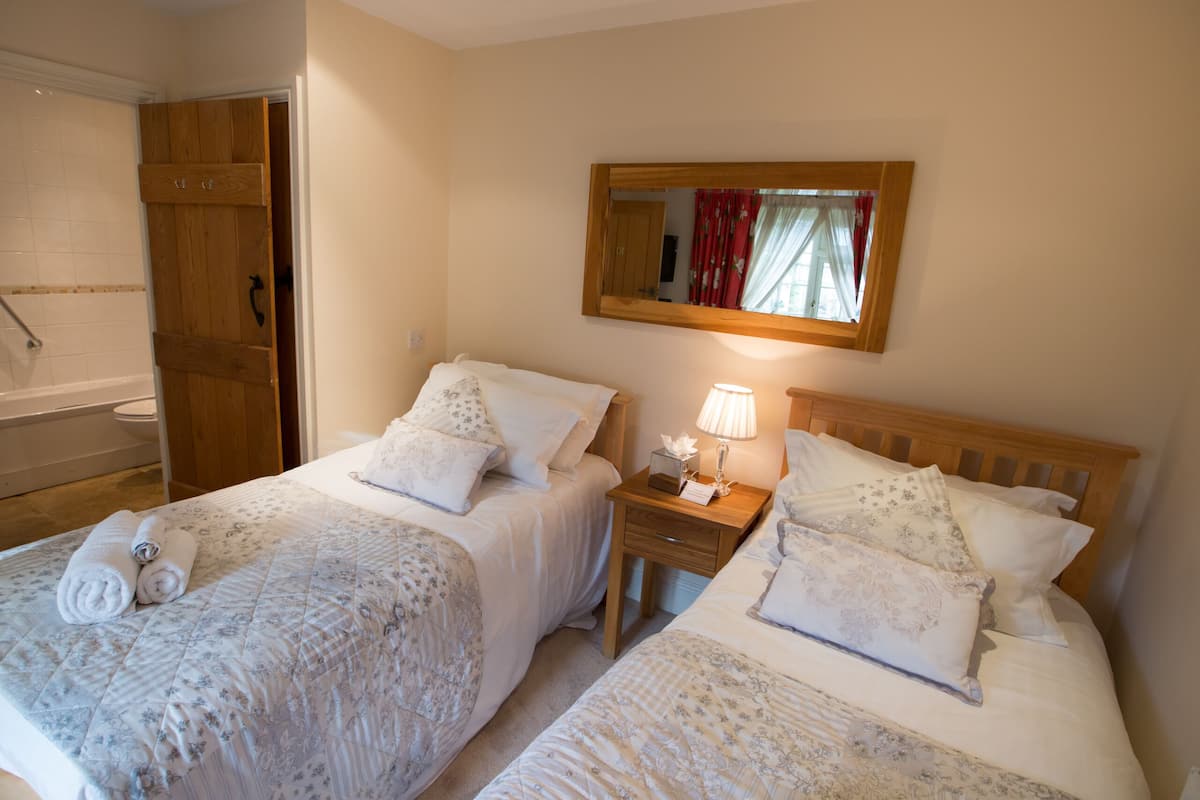 A hotel room with two beds, a wooden wardrobe, a bedside table with a lamp, and a mirror on the wall.
