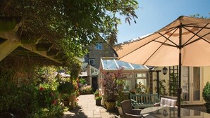 A patio area with a glass table and wicker chairs, surrounded by potted plants and a large umbrella. - The Old Brewhouse (Cirencester)