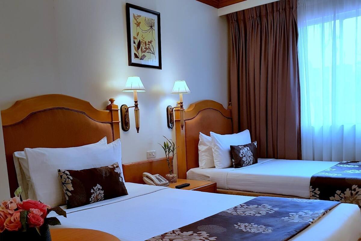A hotel room with two beds, a nightstand with a lamp, a wall-mounted telephone, and a framed picture.