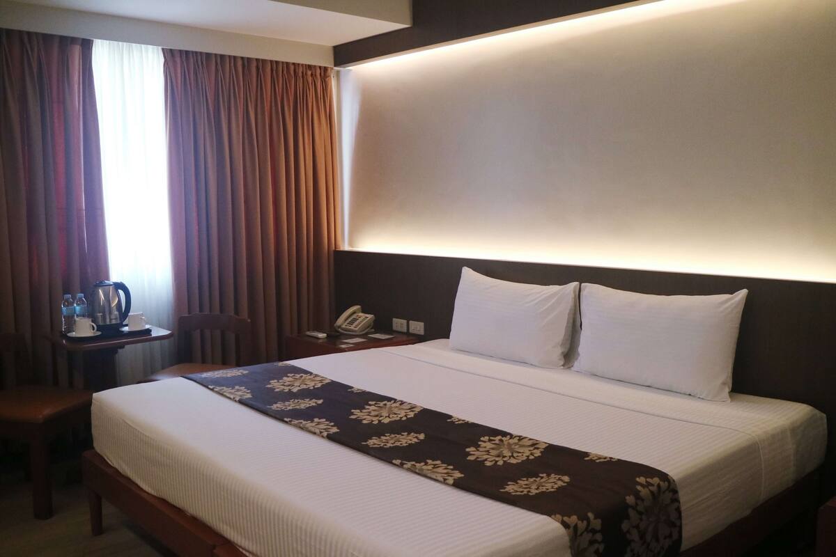 A hotel room with a large bed, a bedside table with a phone, a small table with a kettle and cups, and a window with curtains.