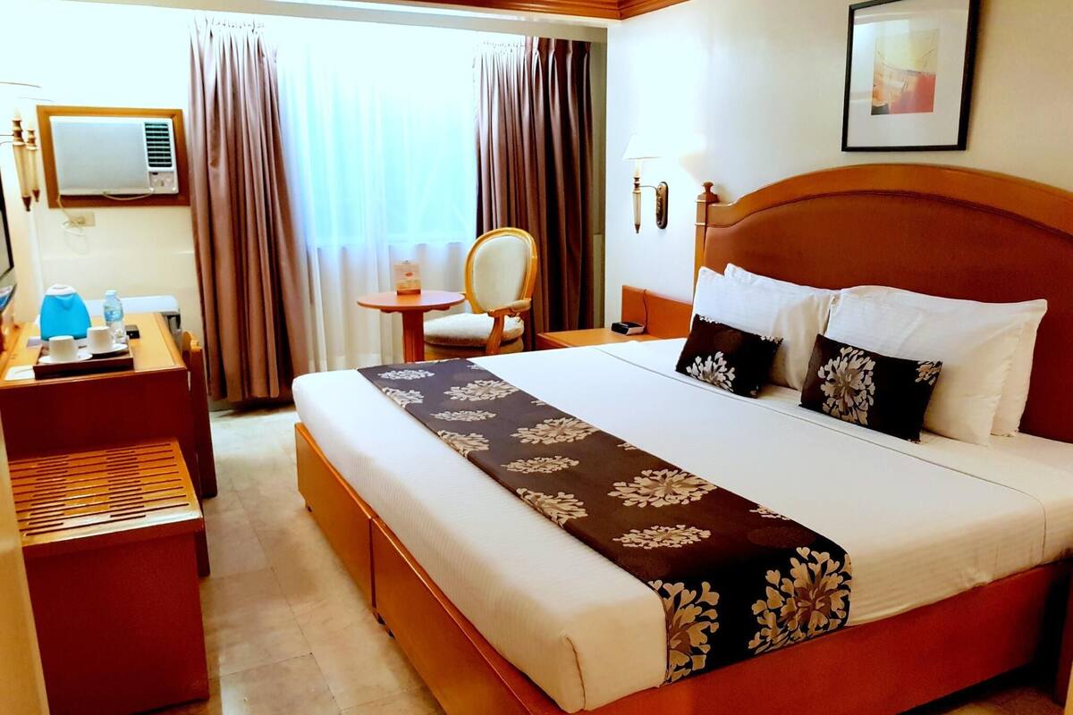 A hotel room with a large bed, a desk, a chair, a small table, and a window with curtains.