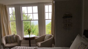 A hotel room with a large bed, two armchairs, a small table with a vase of flowers, and a view of trees through large windows. - Borthalan Hotel (St Ives)