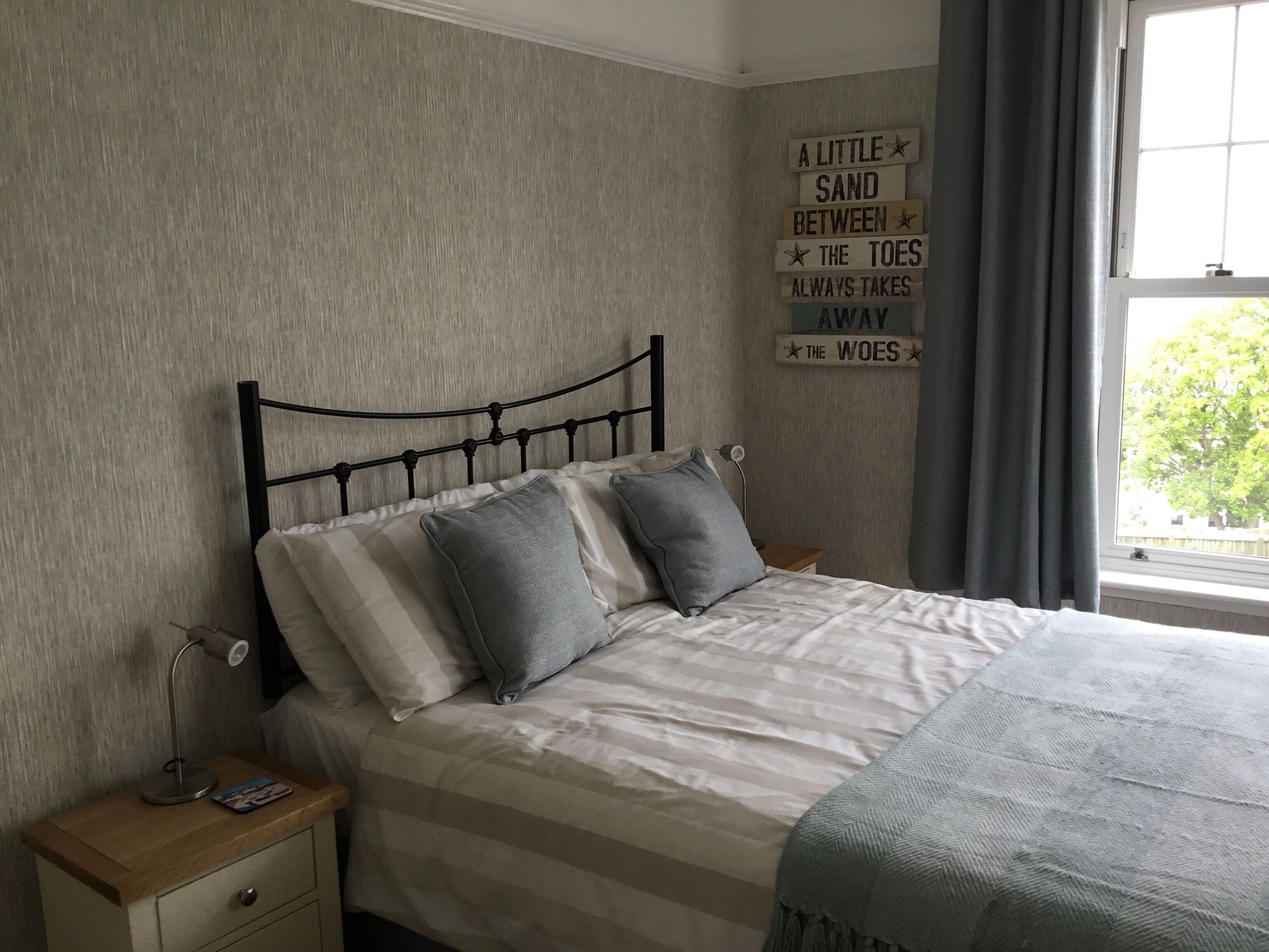 A bedroom with a bed, pillows, a bedside table with a lamp, a wall with a motivational quote, and a window with curtains.
