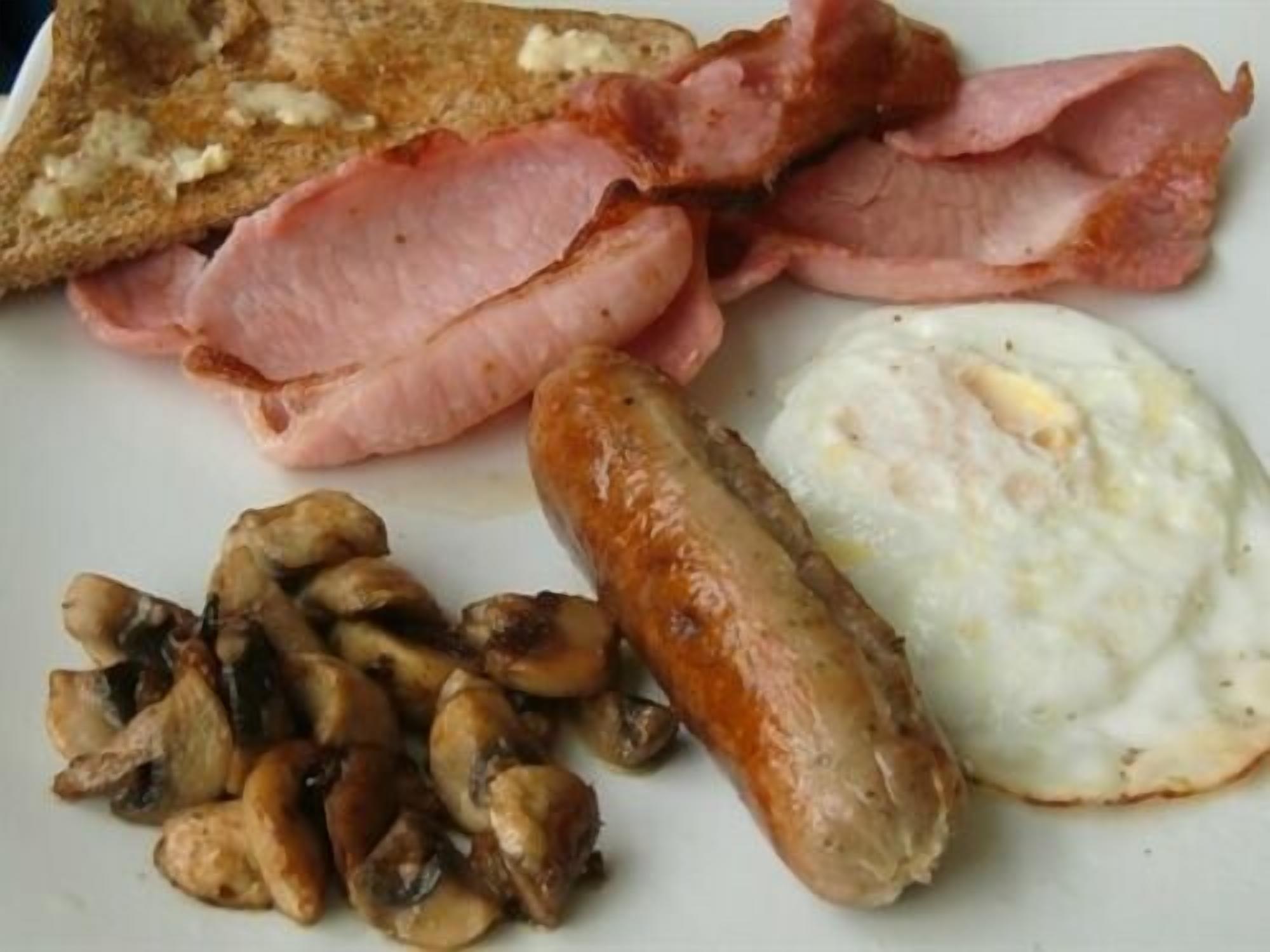 A plate of breakfast food including bacon, sausages, mushrooms, toast, and eggs.
