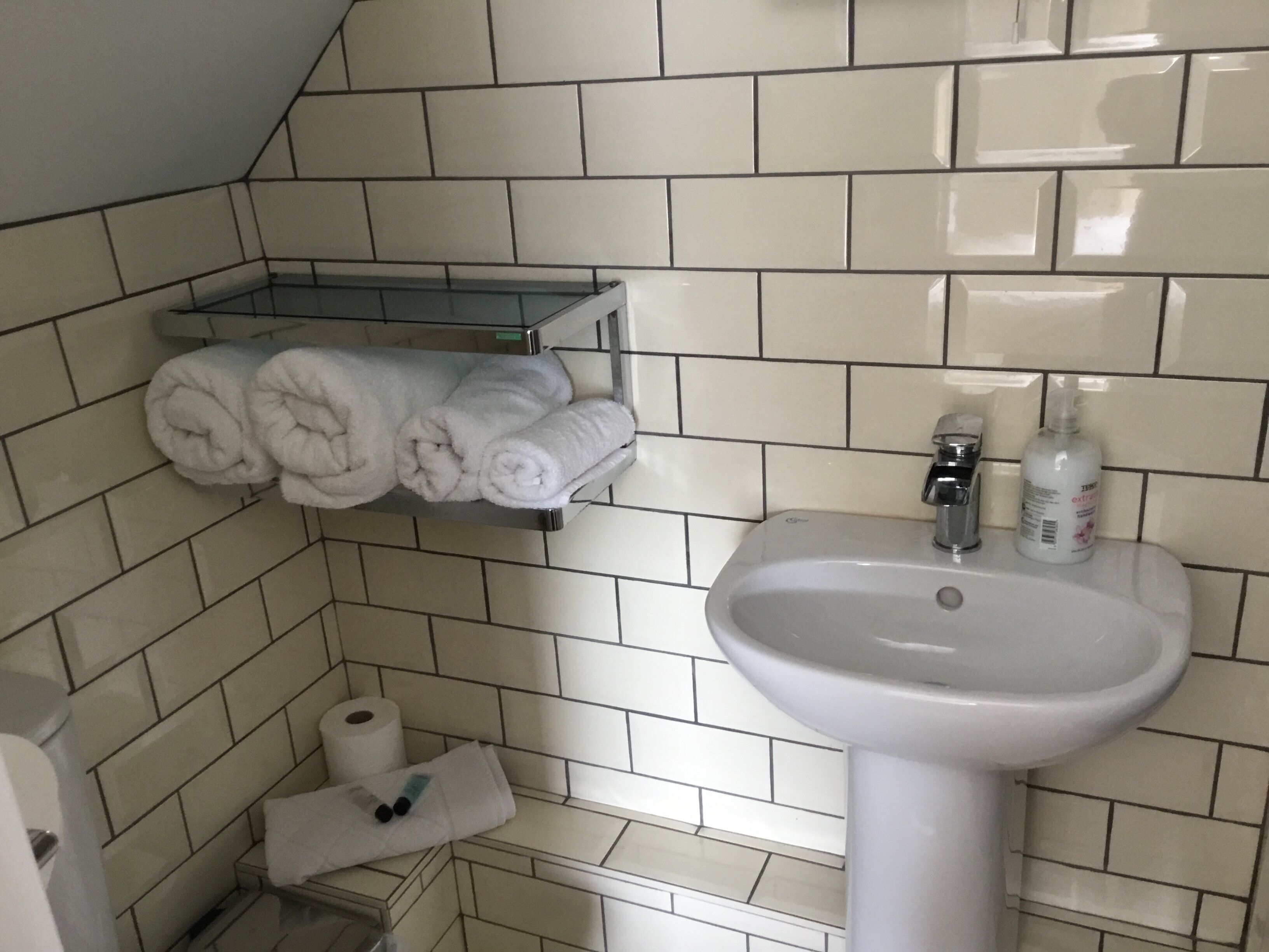 Une salle de bain avec un lavabo blanc, un porte-serviettes avec des serviettes pliées, un rouleau de papier toilette et un flacon de savon pour les mains.