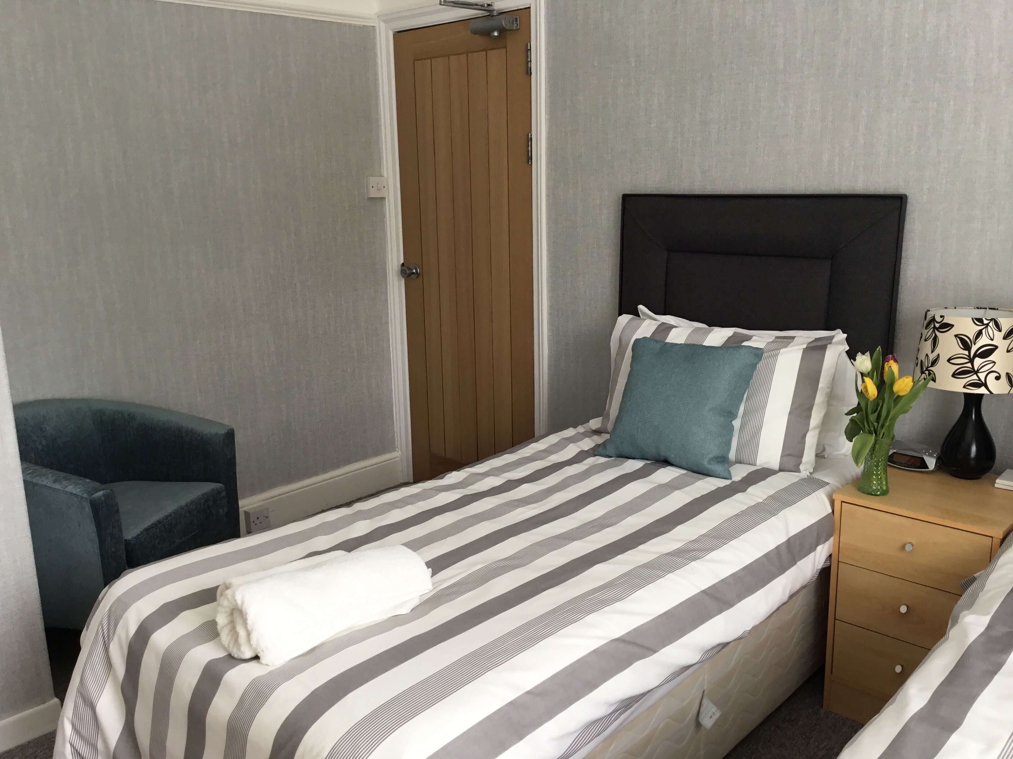 Een net opgemaakt bed met strepenbeddengoed, een nachtkastje met een lamp, een stoel en een deur.
