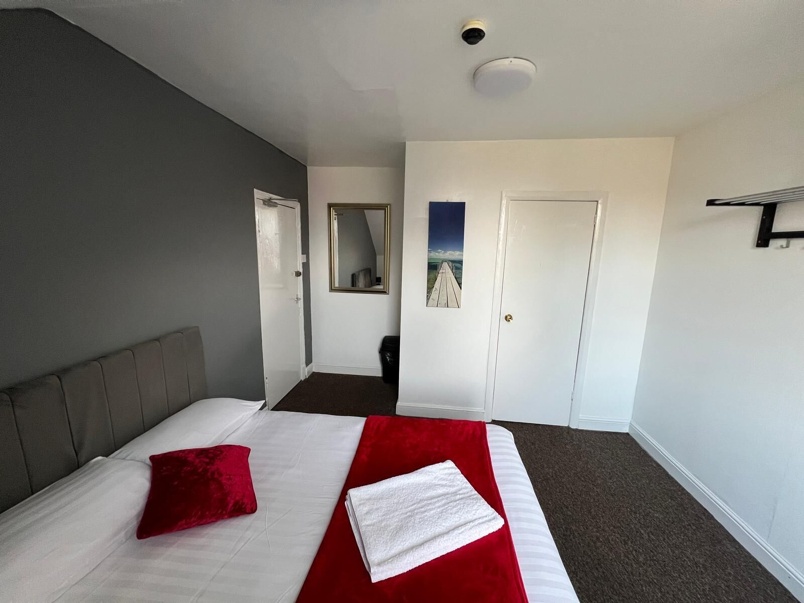 Habitación de hotel con una cama, almohada roja, toalla blanca, cabecera gris, puerta blanca, un espejo y un cuadro en la pared.