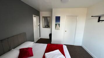 Habitación de hotel con una cama, almohada roja, toalla blanca, cabecera gris, puerta blanca, un espejo y un cuadro en la pared.