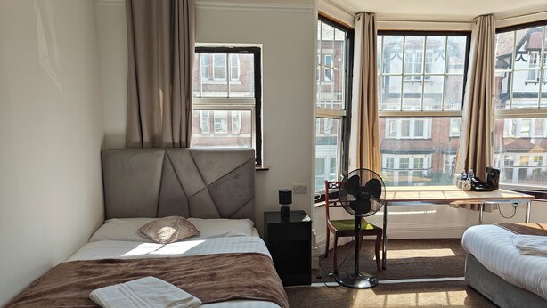 A hotel room with two beds, a desk, a chair, a standing fan, and large windows with curtains. - Campbell's Guest House (Leicester)