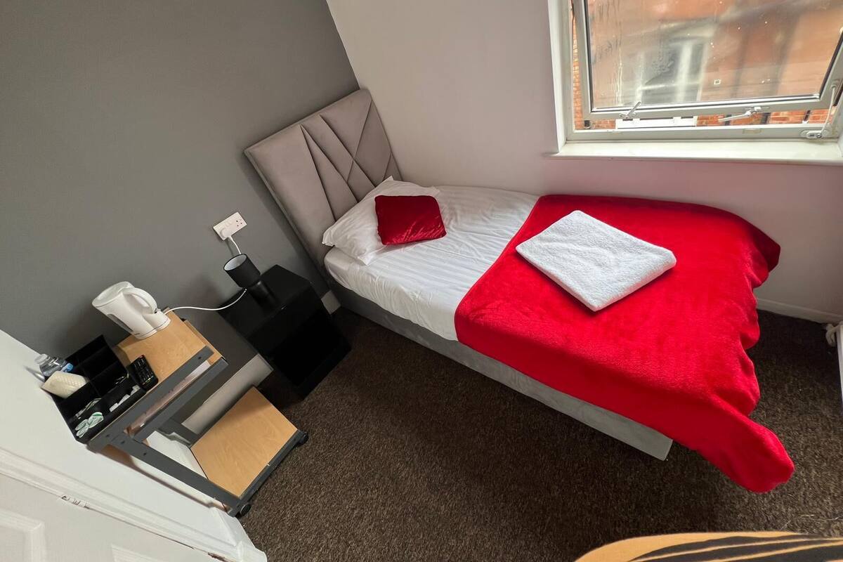 A small, neatly arranged bedroom with a single bed, a red and white bedspread, a white towel, a nightstand with a lamp, a small table with a cup, and a window with a view of buildings.