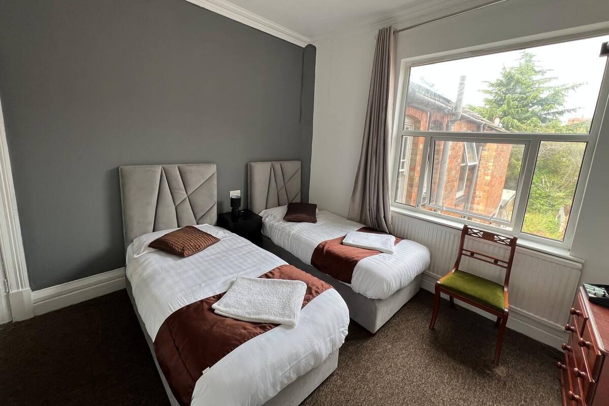 A hotel room with two beds, a chair, a radiator, and a window with a view of buildings.