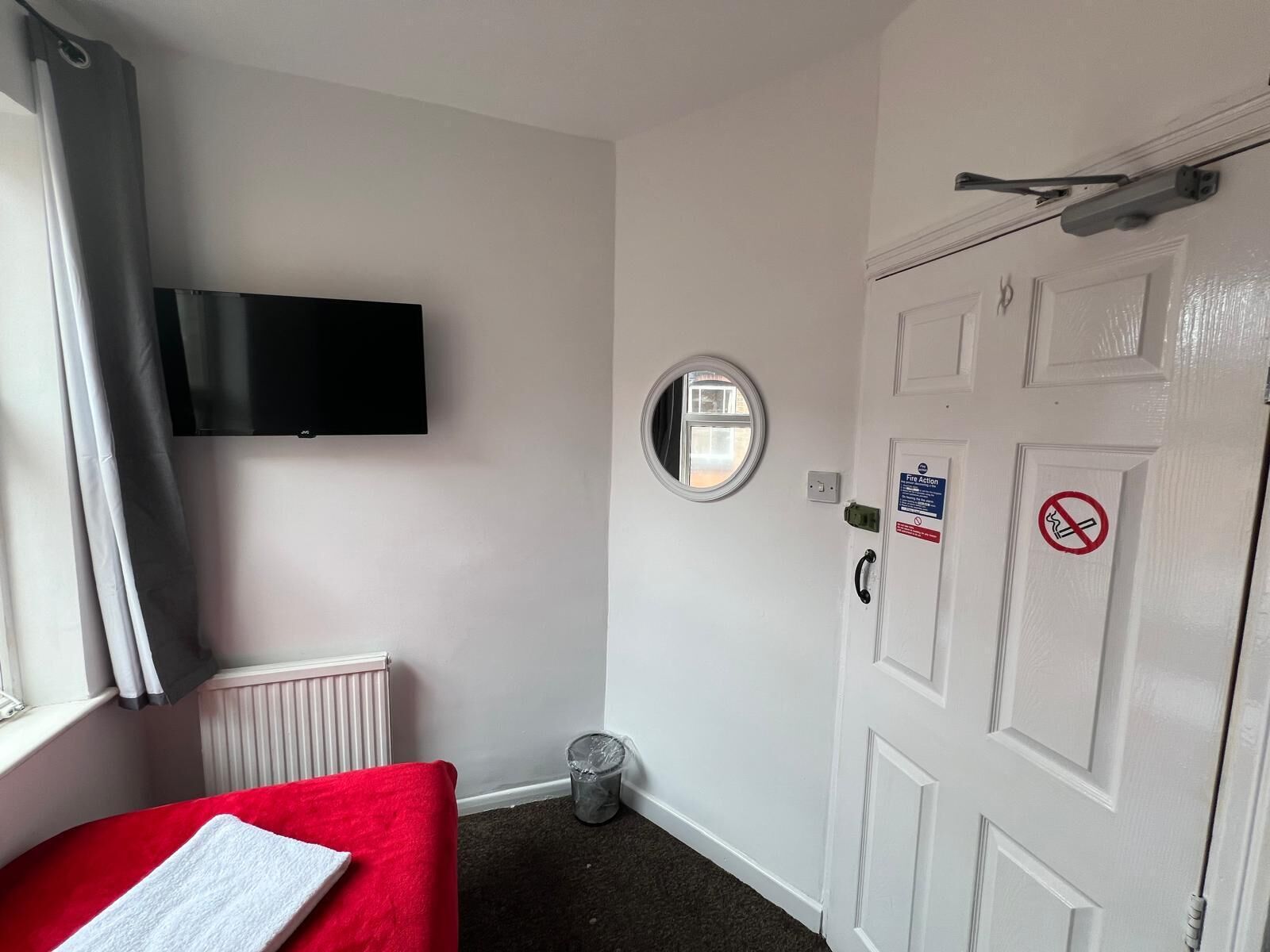 Hotelkamer met een bed, een aan de muur bevestigde televisie, een deur met een pictogram voor brandveiligheid, een raam met gordijnen, en een radiator.