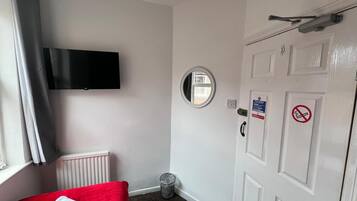 Hotelkamer met een bed, een aan de muur bevestigde televisie, een deur met een pictogram voor brandveiligheid, een raam met gordijnen, en een radiator.