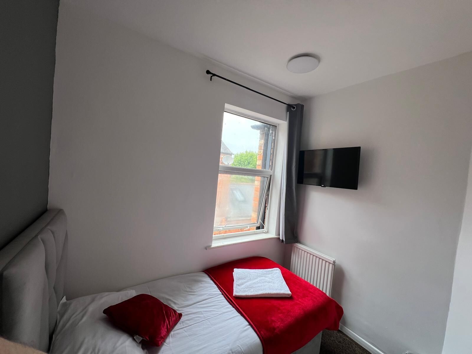 A small, single-bed room with a red bedspread, a white pillow, and a red pillow on the bed. There is a window with a view of a brick building outside. A flat-screen TV is mounted on the wall above the bed.