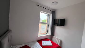 A small, single-bed room with a red bedspread, a white pillow, and a red pillow on the bed. There is a window with a view of a brick building outside. A flat-screen TV is mounted on the wall above the bed.