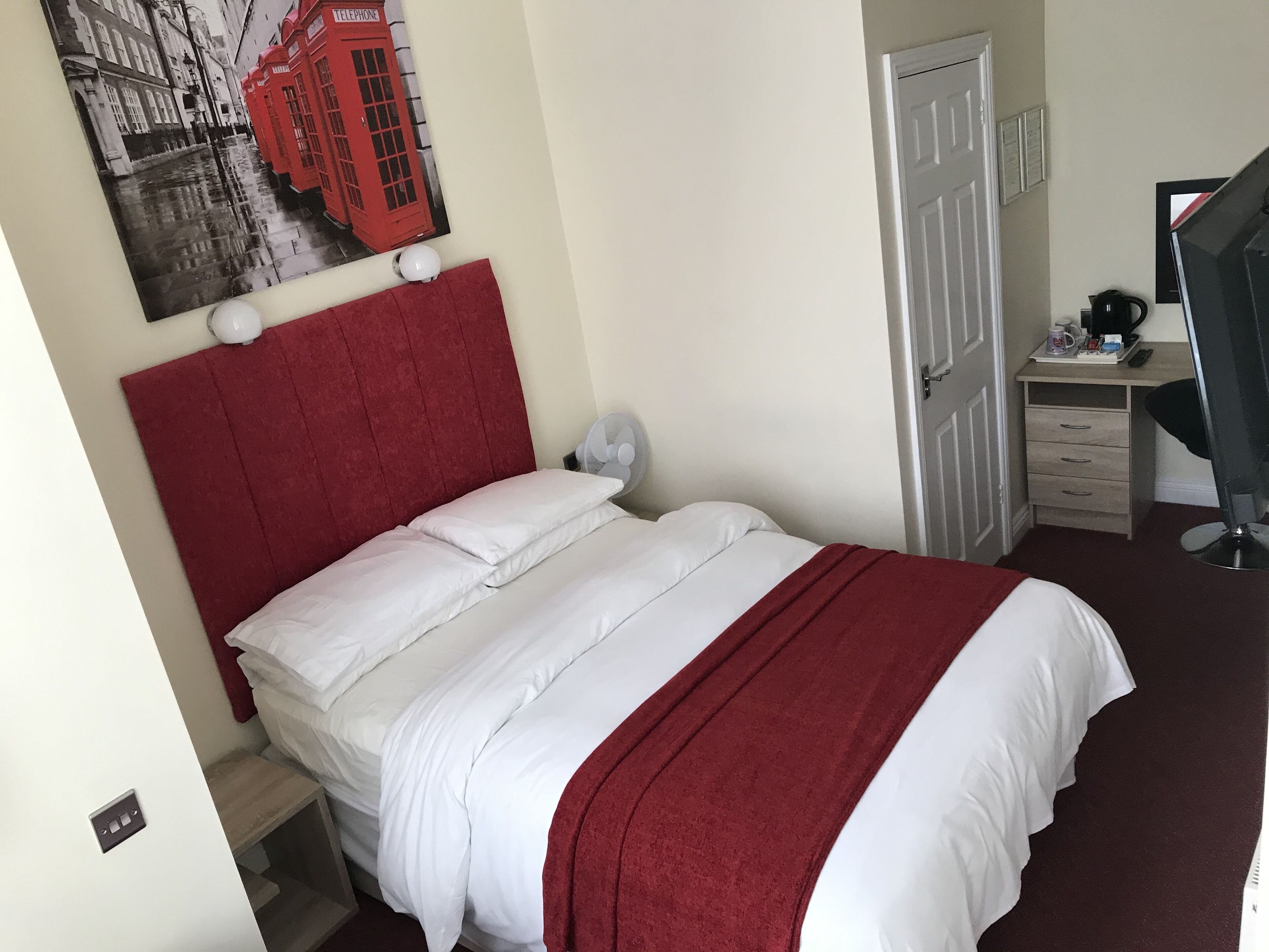 A neatly made bed with white bedding and a red headboard, a desk with a computer and a coffee maker, and a wall-mounted telephone booth artwork.