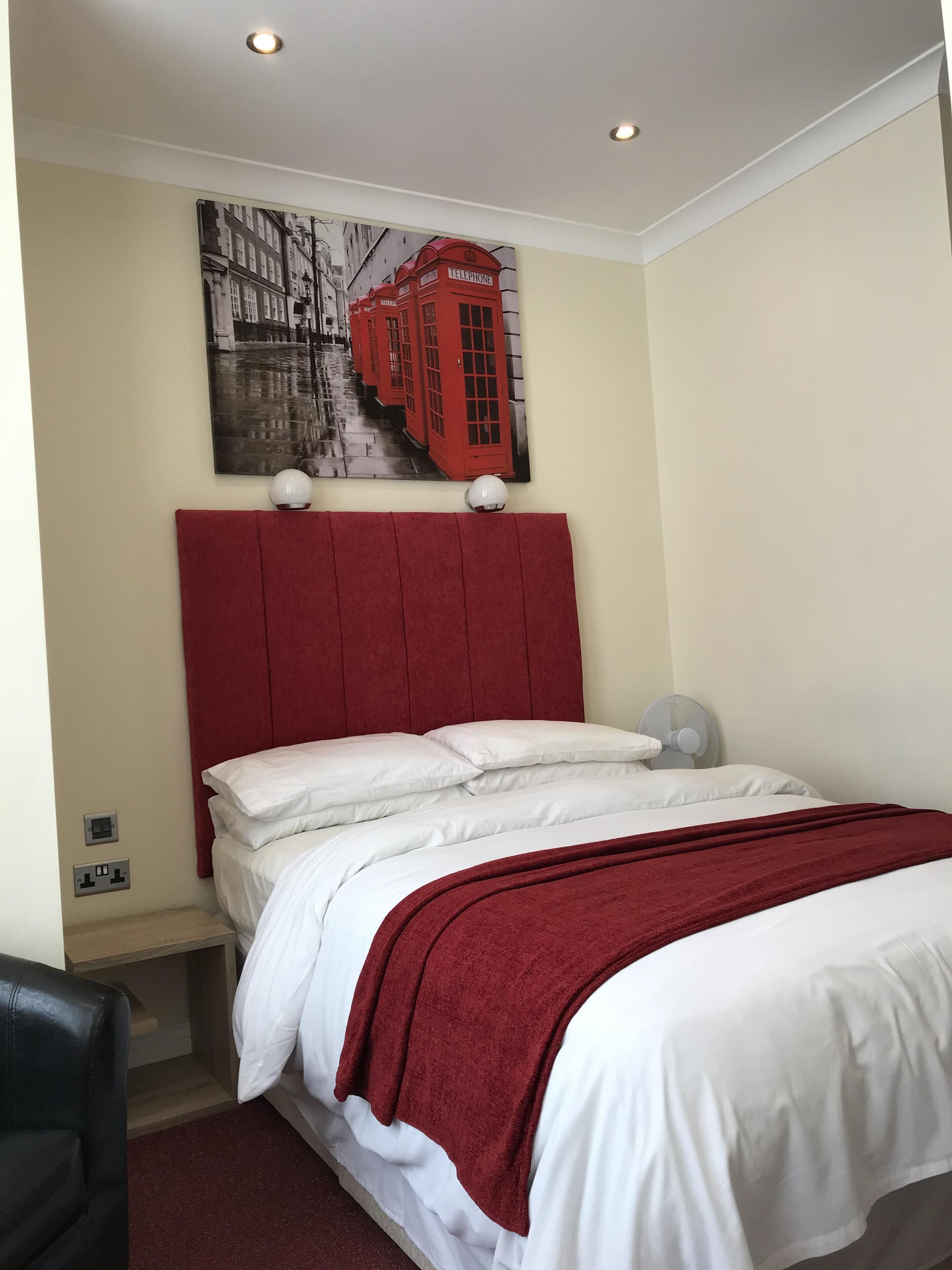 Una habitación de hotel con una cama, cabecera roja, manta blanca, plaid rojo, luz empotrada en la pared, ventilador y un cuadro de un teléfono rojo.