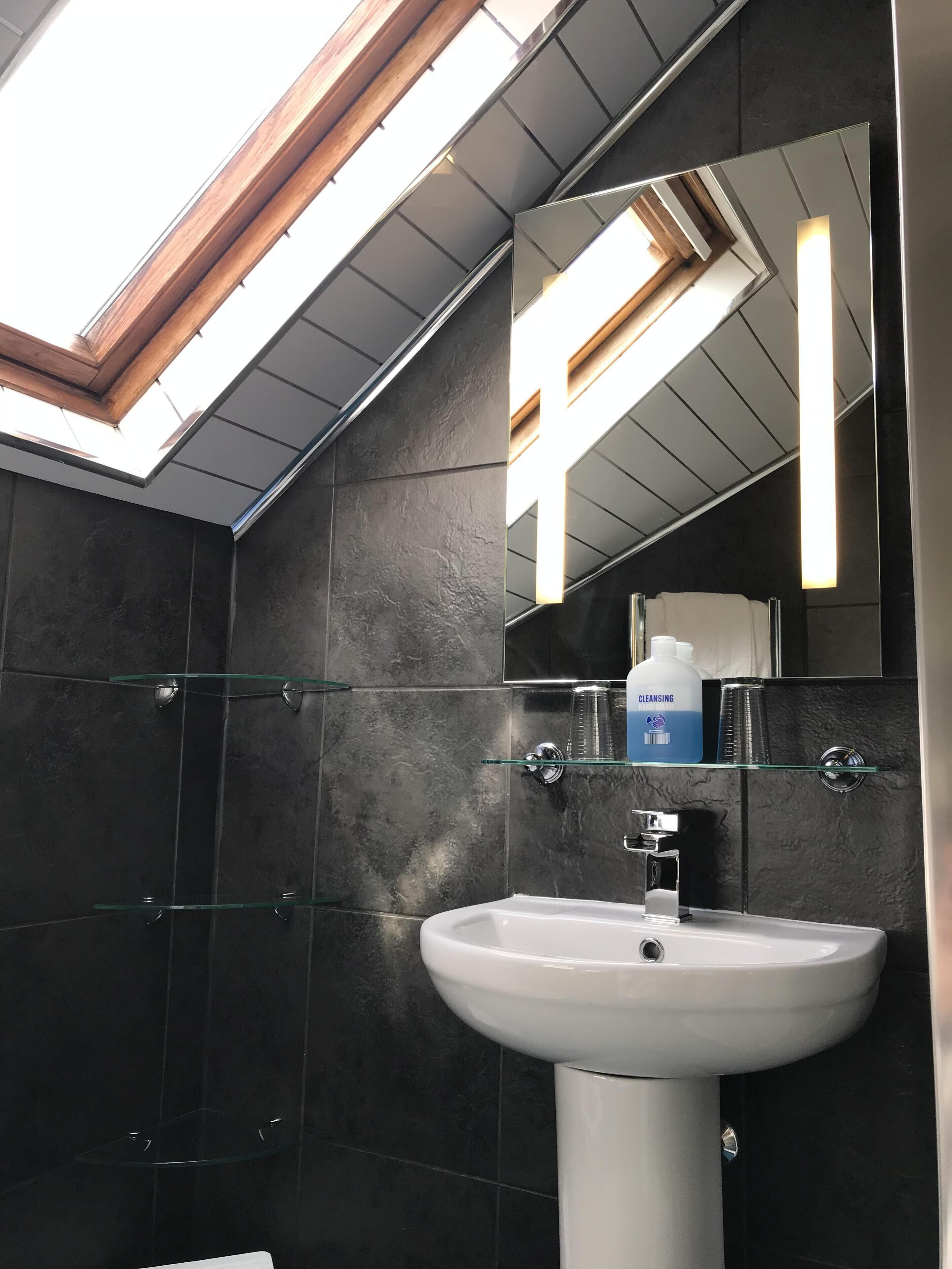 Een moderne badkamer met dakraam, donkere tegels, een glazen plank met toiletartikelen en een witte wastafel.