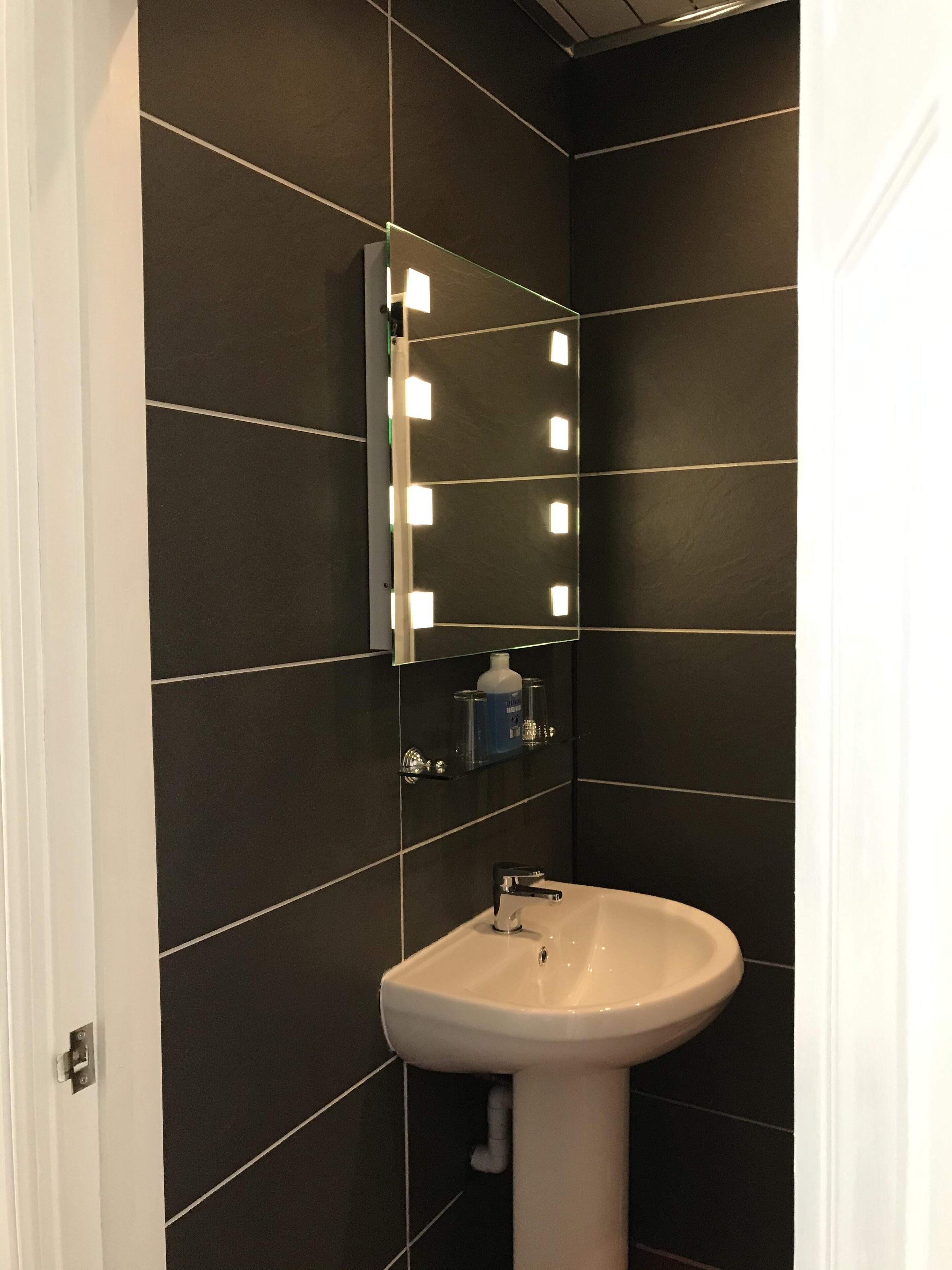 A bathroom with dark tiled walls, a white sink, and a mirror with lights.