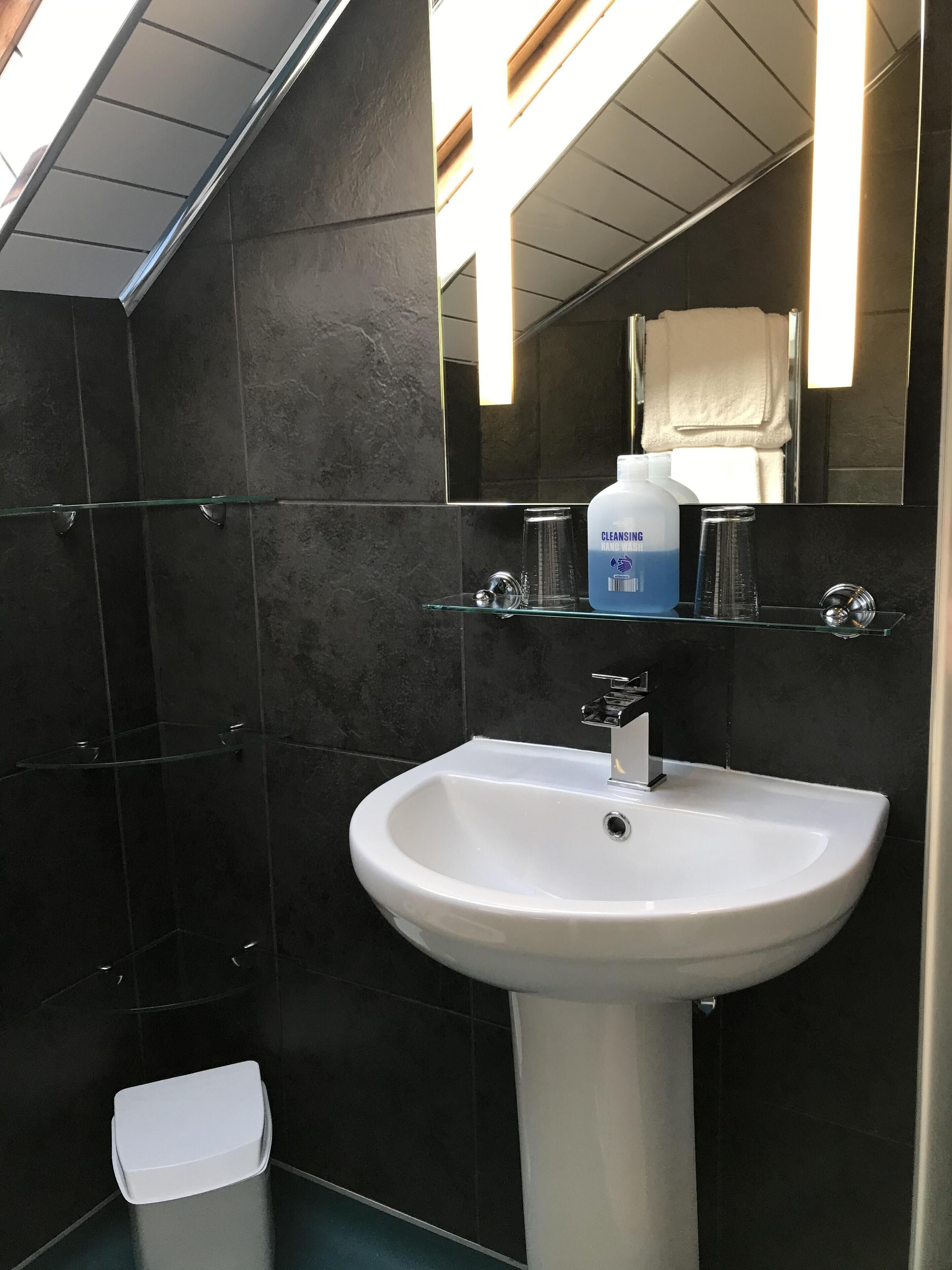 A bathroom with a white sink, dark tiled walls, a glass shelf with toiletries, and a mirror reflecting a towel.