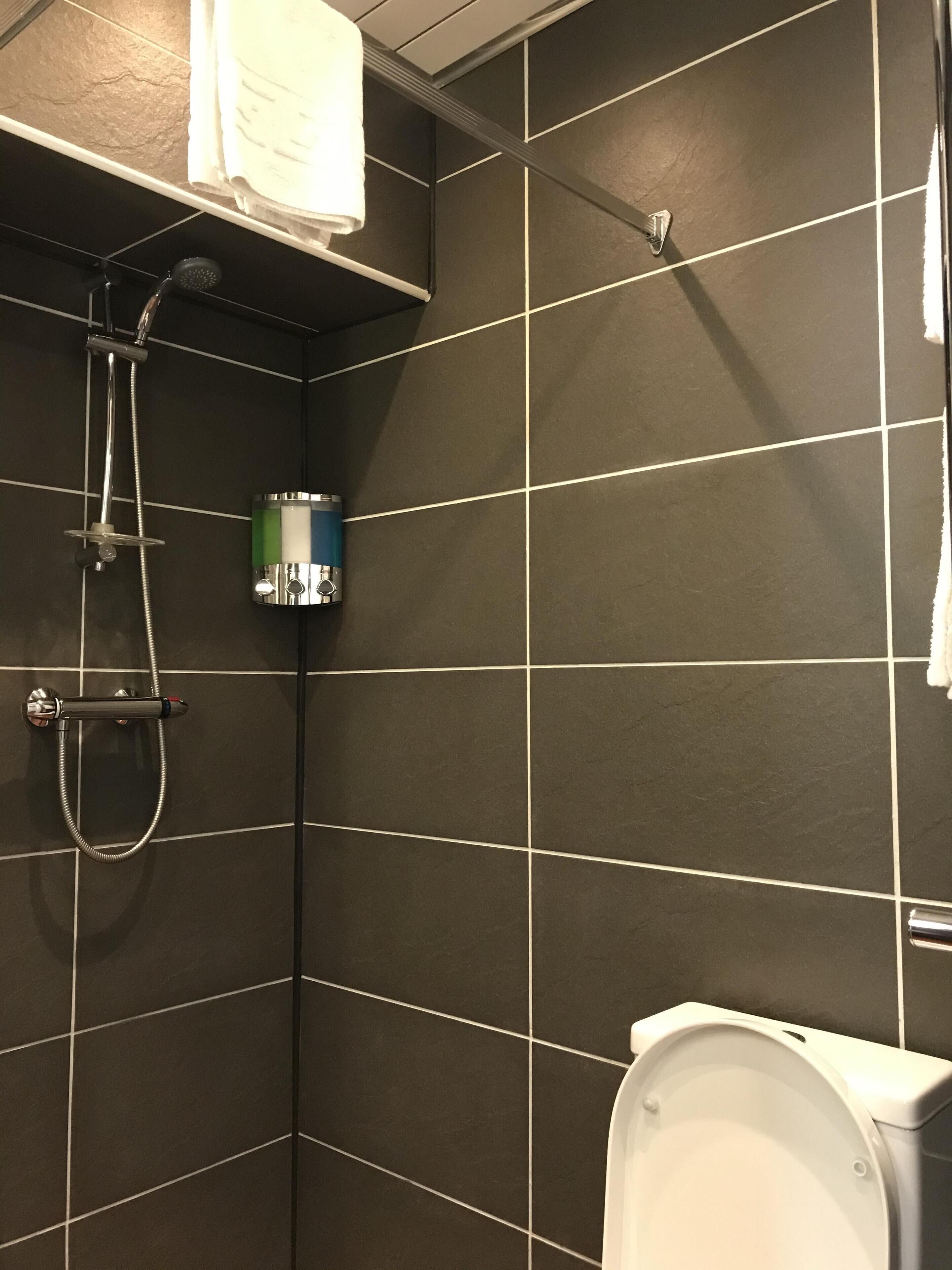 A bathroom with dark tiled walls, a shower with a handheld showerhead, a toilet, and a towel hanging on the wall.