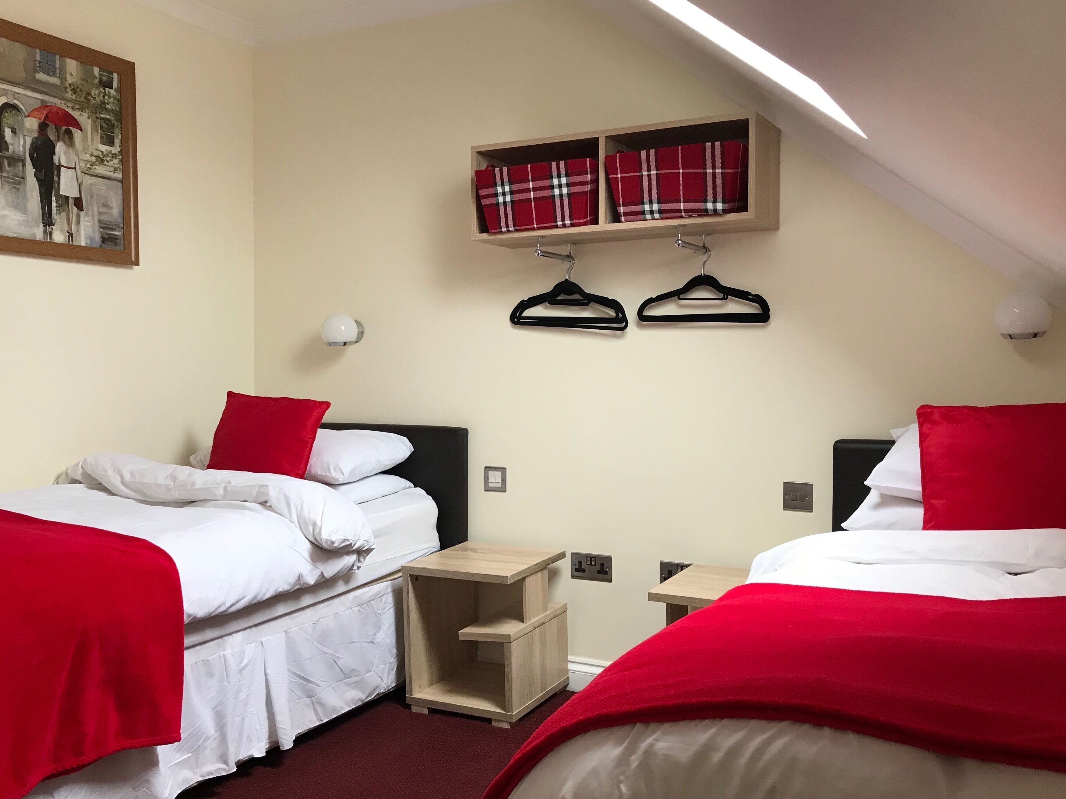 A hotel room with two beds, each with red and white bedding, a small wooden nightstand, a wall-mounted shelf with plaid towels, and a painting of a couple on a bridge.