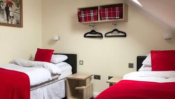 A hotel room with two beds, each with red and white bedding, a small wooden nightstand, a wall-mounted shelf with plaid towels, and a painting of a couple on a bridge.
