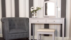 A grey upholstered armchair with decorative studs, a white vanity with a mirror, and a small stool. - Castle Park Hotel (Leicester)