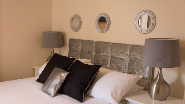 A neatly made bed with a stone headboard, two bedside tables with lamps, and three wall mirrors. - Castle Park Hotel (Leicester)