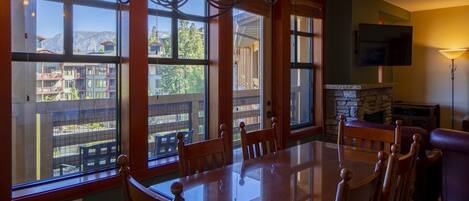 Comedor con una gran mesa de vidrio y sillas de madera, ventanas amplias con vistas a edificios y montañas, chimenea de piedra y un televisor de pantalla plana montado.