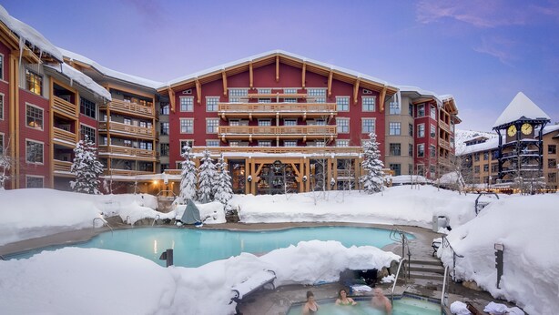 Un hotel con piscina rodeado de nieve, balcones de madera y un gran edificio al fondo.