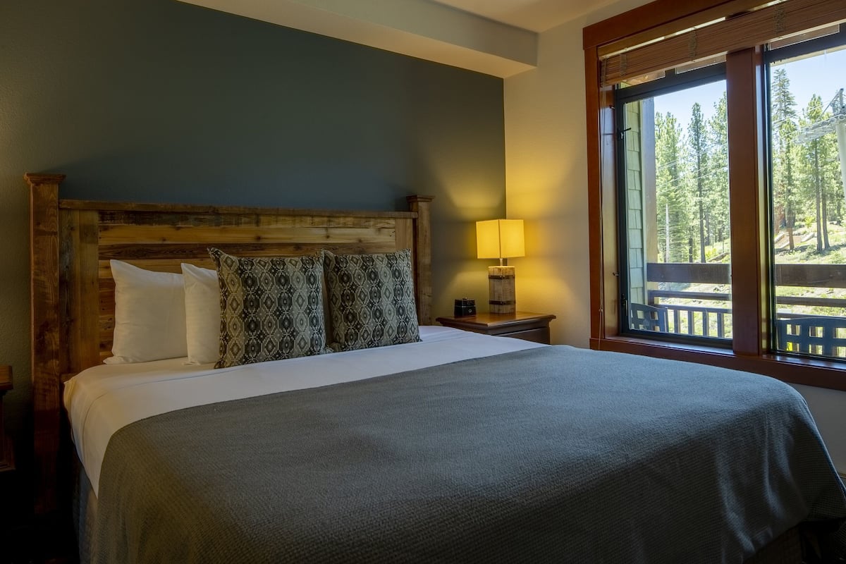 A bed with a wooden headboard, white and patterned pillows, and a grey bedspread. There is a nightstand with a lamp and a view of a deck and trees through a window.