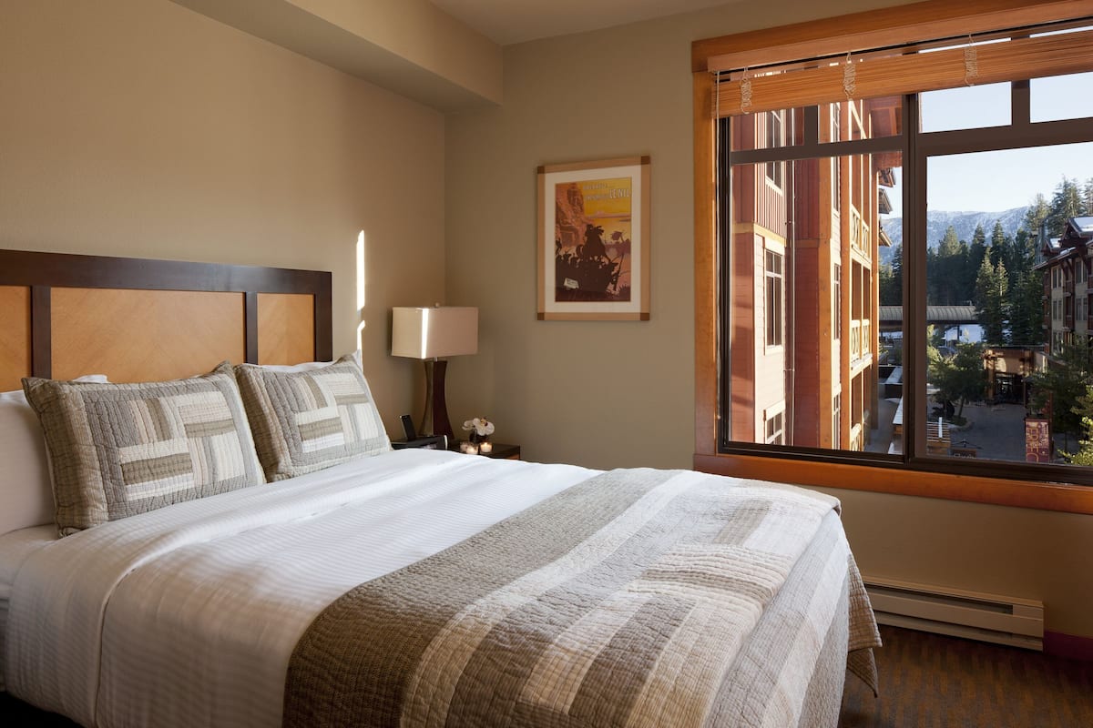 A neatly made bed with a striped blanket and pillows, a bedside table with a lamp, a framed picture on the wall, and a window with a view of buildings and trees.