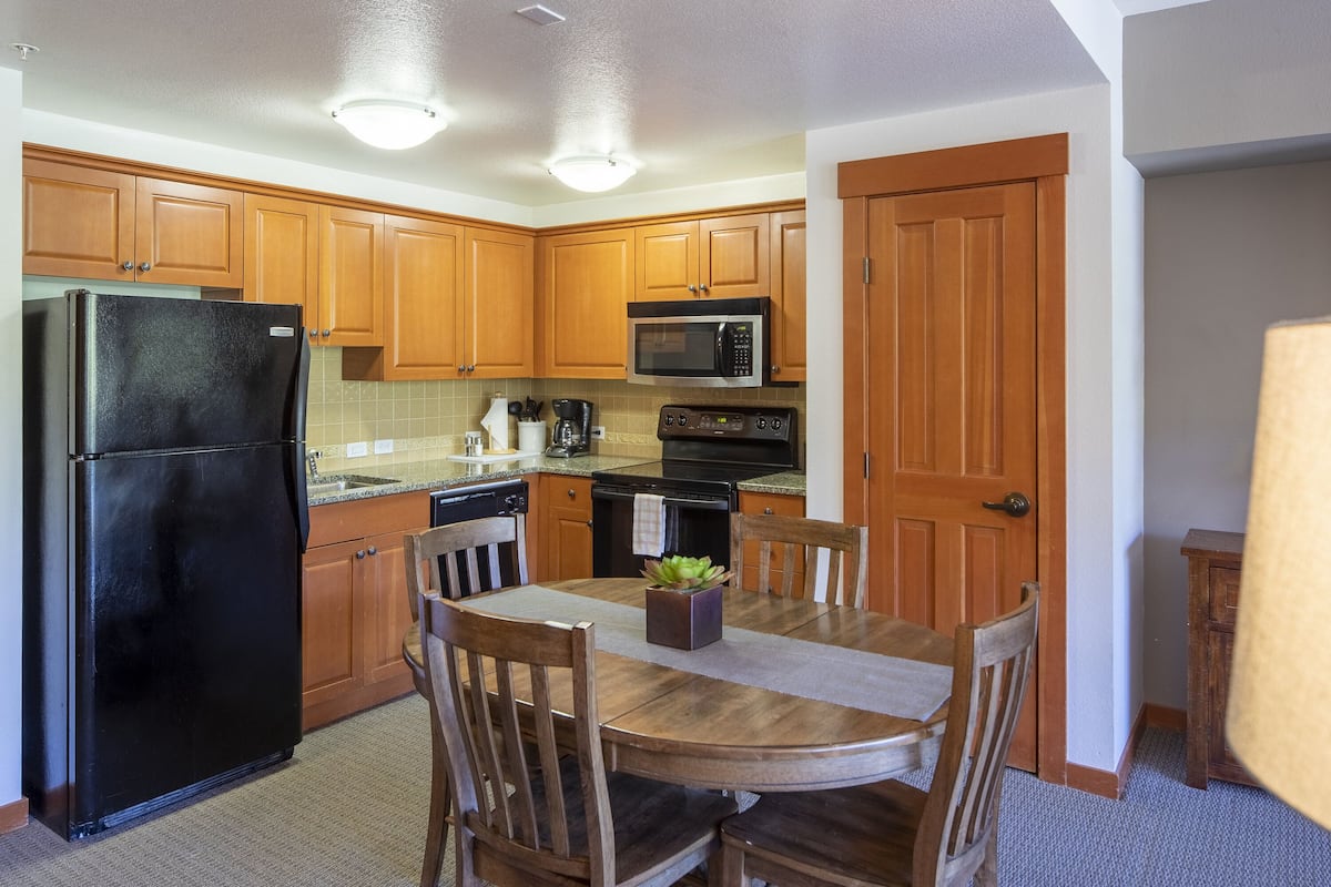 A compact kitchen with wooden cabinets, a black refrigerator, a microwave, and a stove. There is a dining table with chairs and a small plant on it.