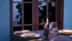 Une table à manger dressée avec des assiettes, des couverts et des verres, placée près d’une fenêtre donnant sur la mer.