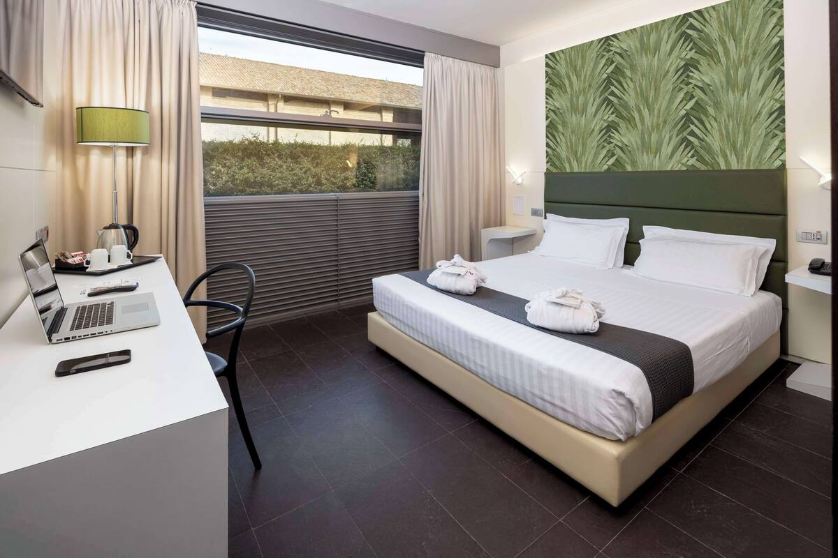 A hotel room with a bed, desk, laptop, and a view through the window.