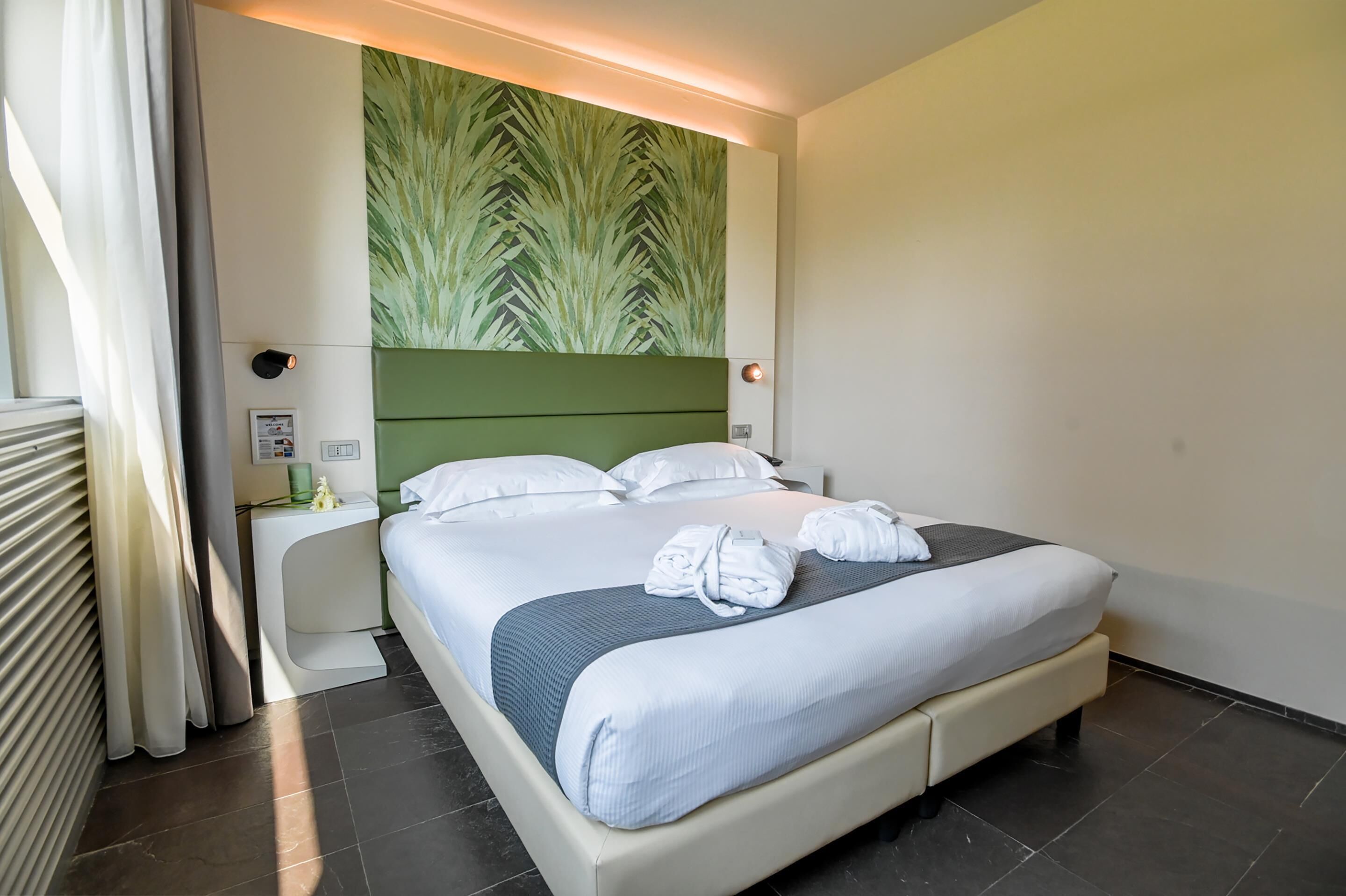 Een hotelkamer met een bed, een nachtkastje, een wandlamp en een groene muur met bladmotief.