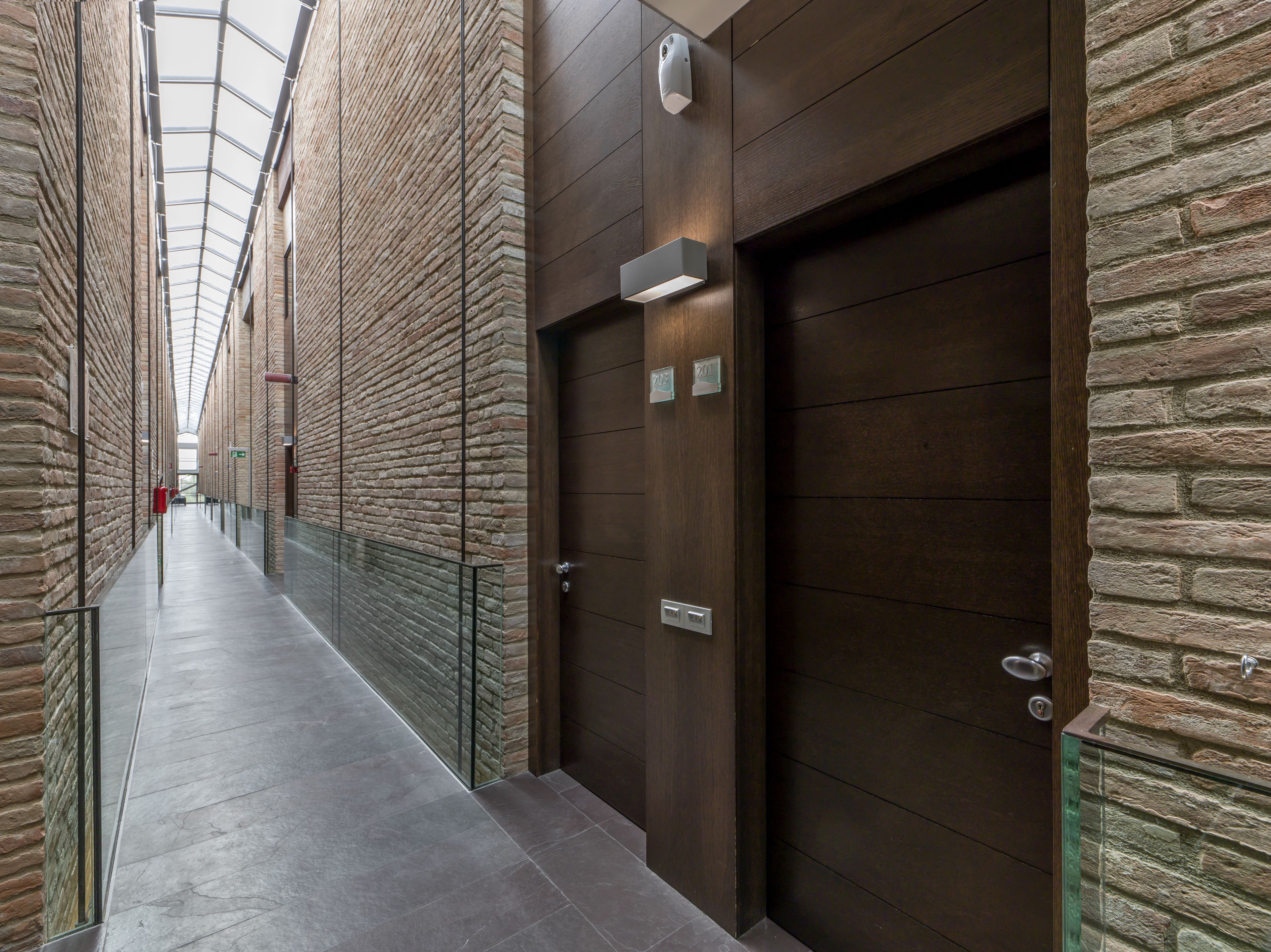 A corridor with stone walls, a glass railing, and a door with a number plate.
