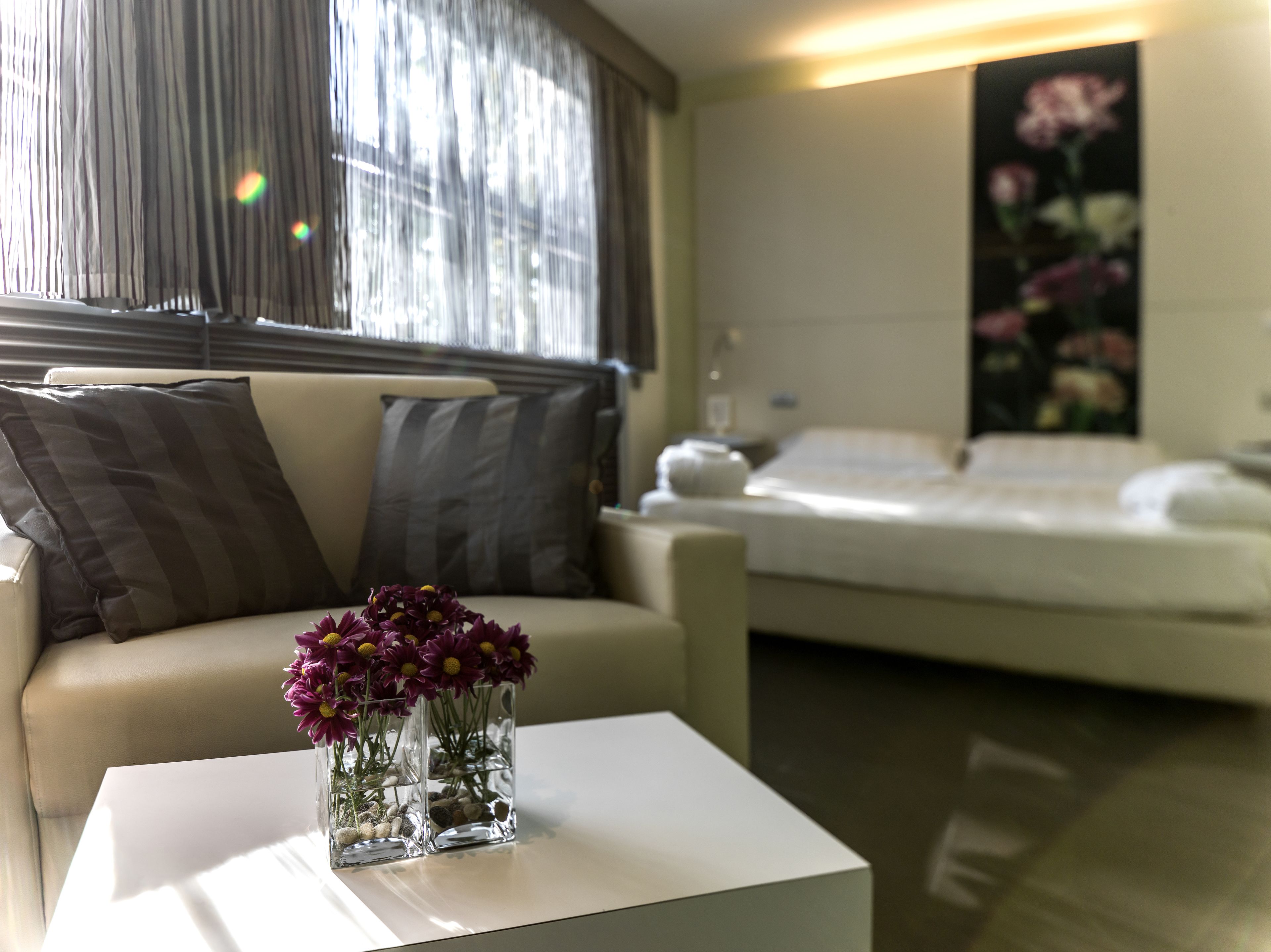 A modern living room with a sofa, a coffee table with a vase of flowers, and a bed in the background.