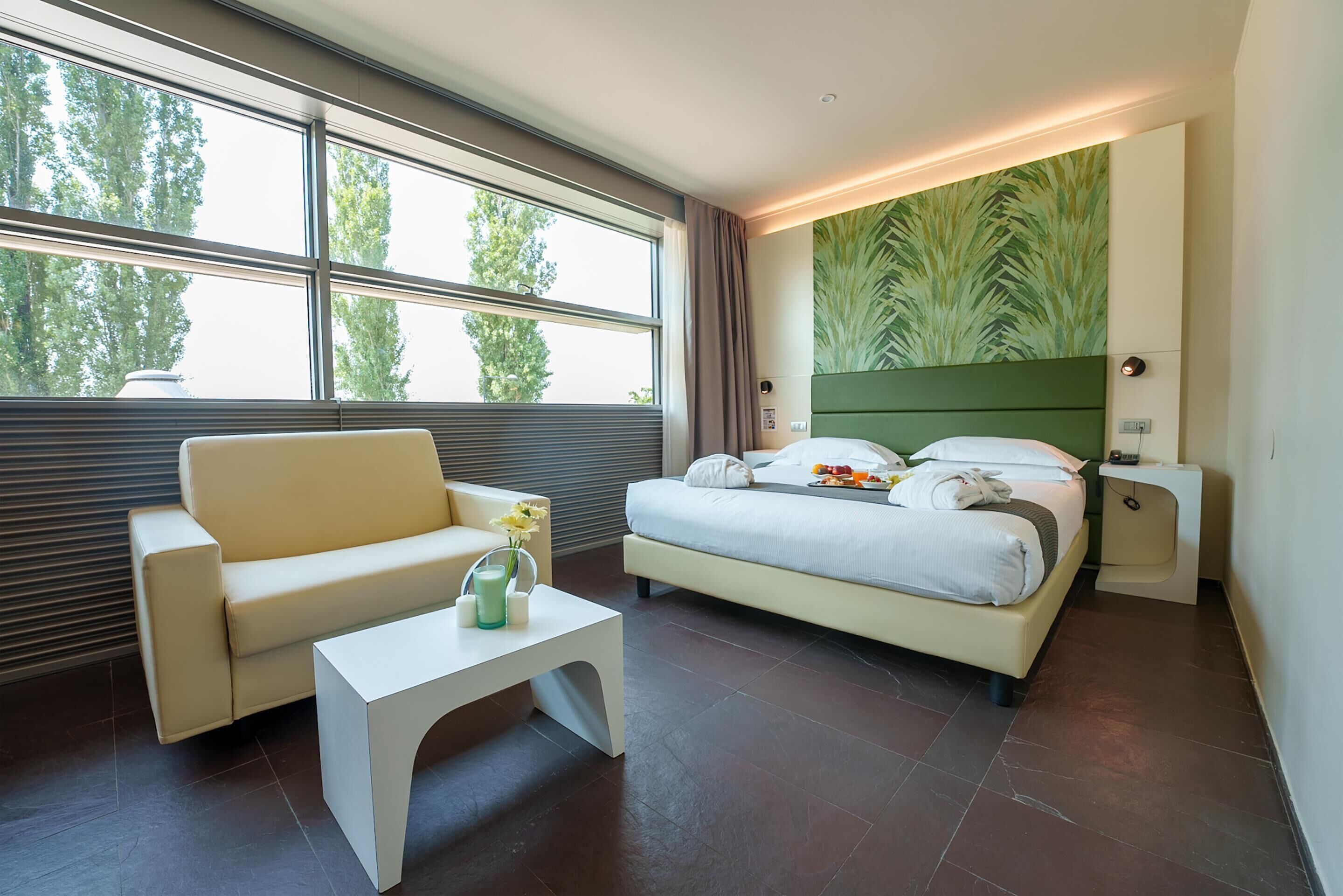 A modern hotel room with a large window, a bed with a green headboard, a white sofa, and a small white table.
