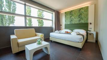 A modern hotel room with a large window, a bed with a green headboard, a white sofa, and a small white table.